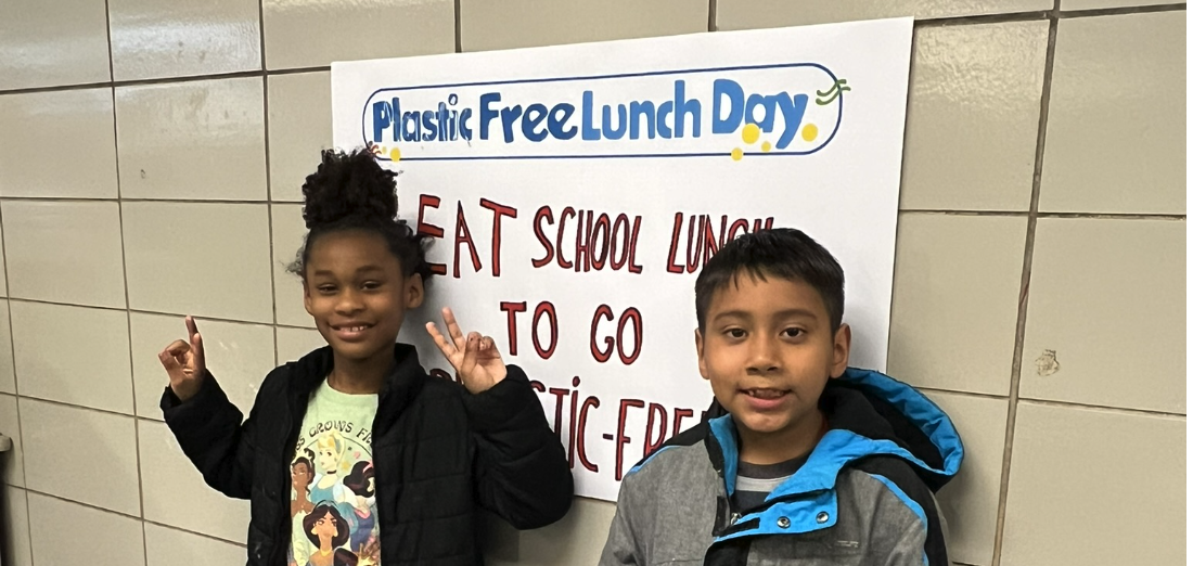 Two children stand in front of a sign that reads “Plastic Free Lunch Day” and encourages eating school lunch to go plastic-free.
