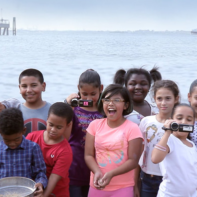 A group of children stand together by the water, some holding video cameras, with a pier visible in the background.