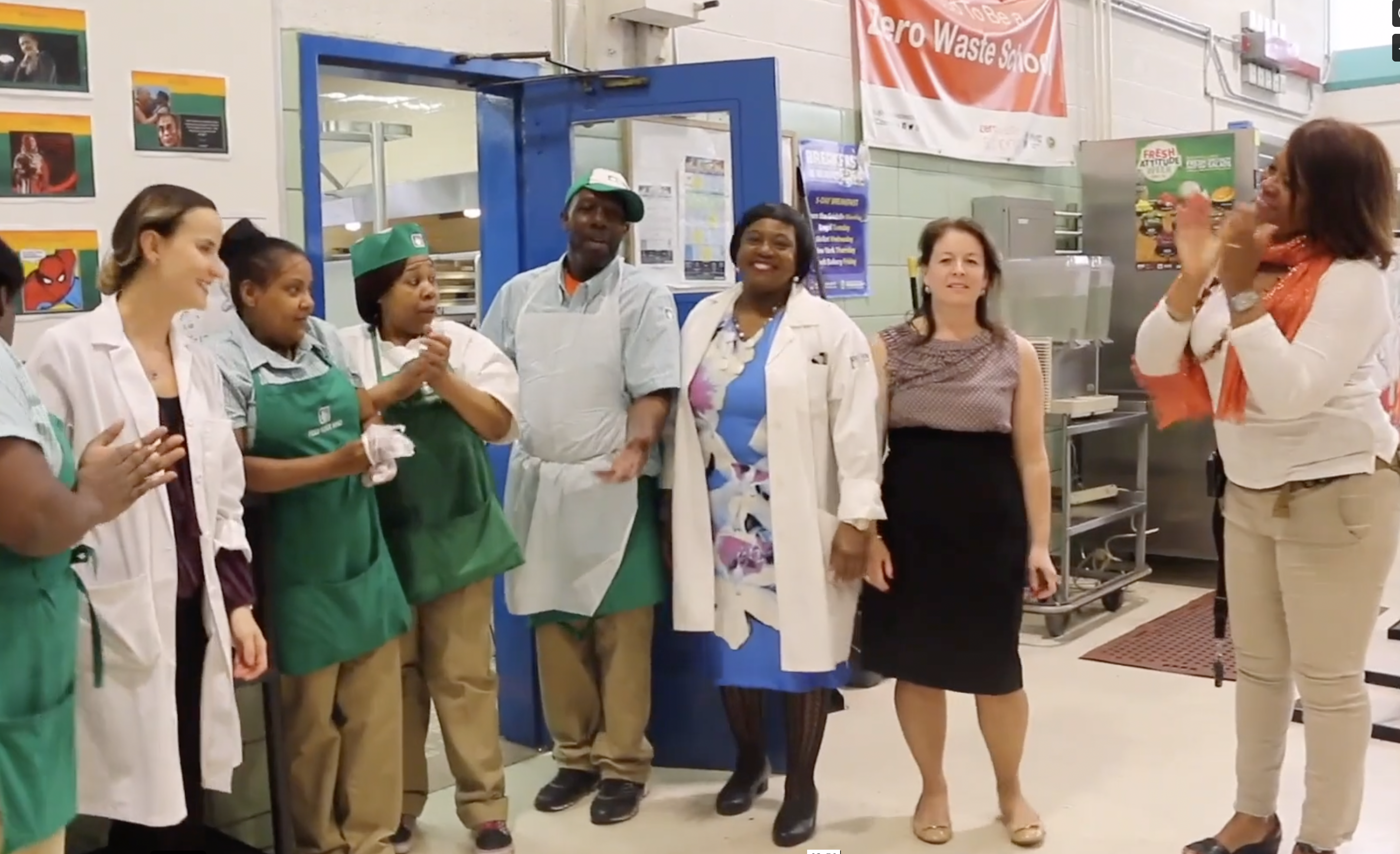 A group of people, including kitchen staff in uniforms and others in business attire, stand together in a commercial kitchen near a blue door while one person applauds.