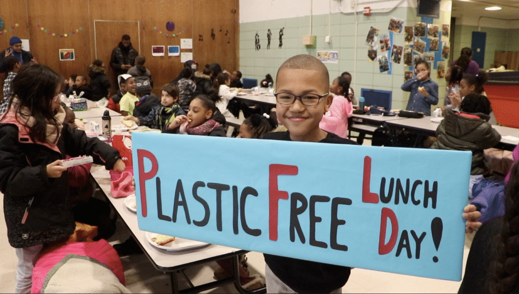 A child holds a “Plastic Free Lunch Day” sign in a crowded school cafeteria with students eating lunch in the background.