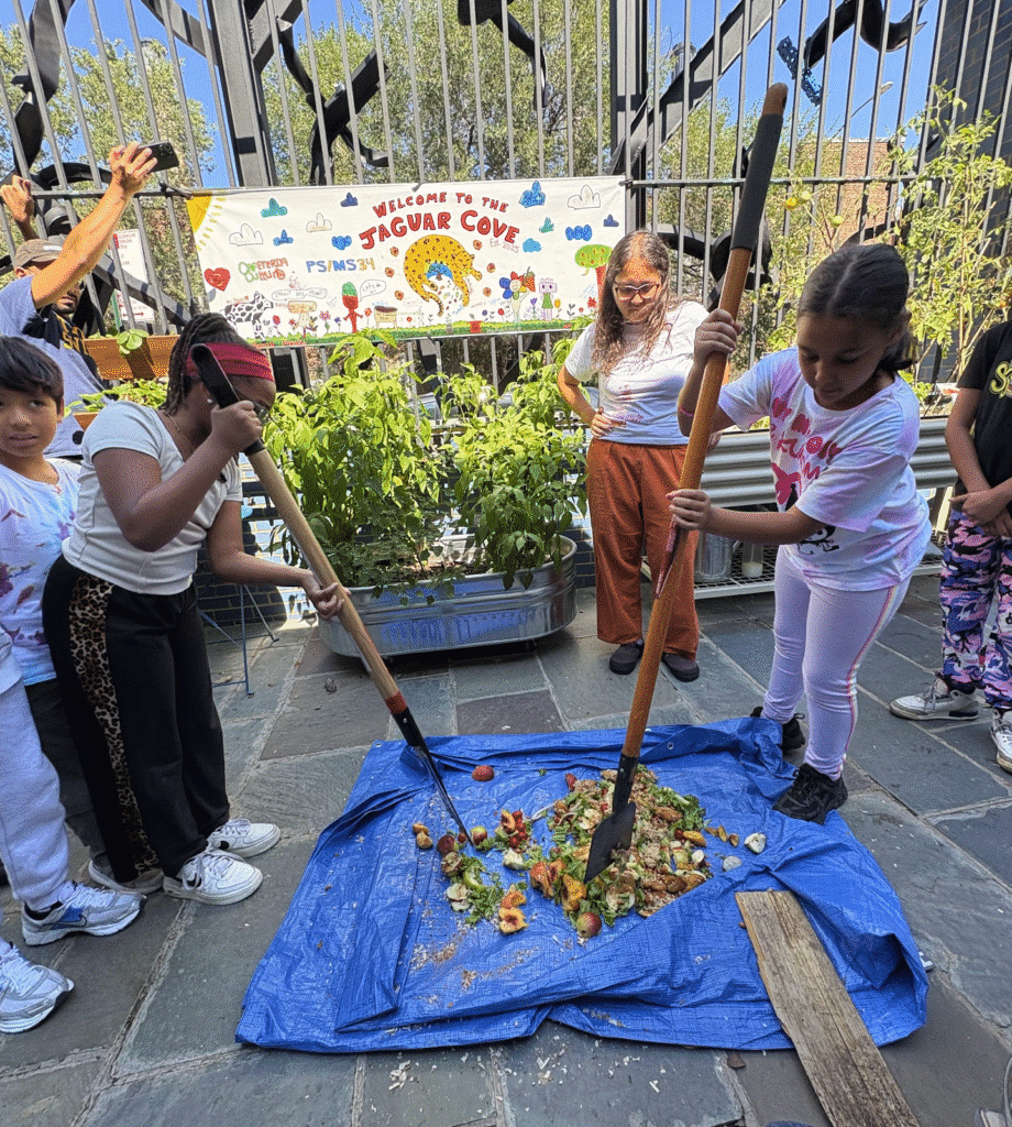 Children use shovels to mix food scraps on a blue tarp outdoors, with a garden, plants, and a "Jaguar Cove" sign in the background.