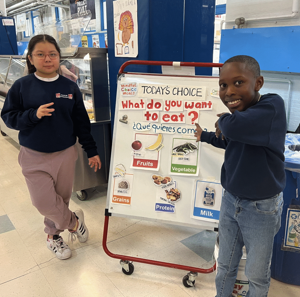 Two children stand beside a cafeteria menu board displaying food choices: fruits, vegetables, grains, protein, and milk, with text in English and Spanish.
