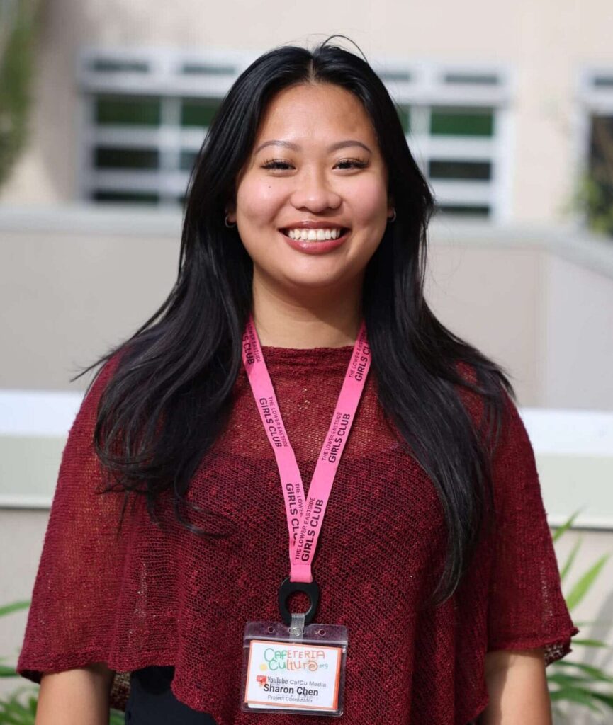A woman with long black hair, wearing a red top and pink lanyard with a name badge, stands outdoors in front of a building, smiling at the camera.