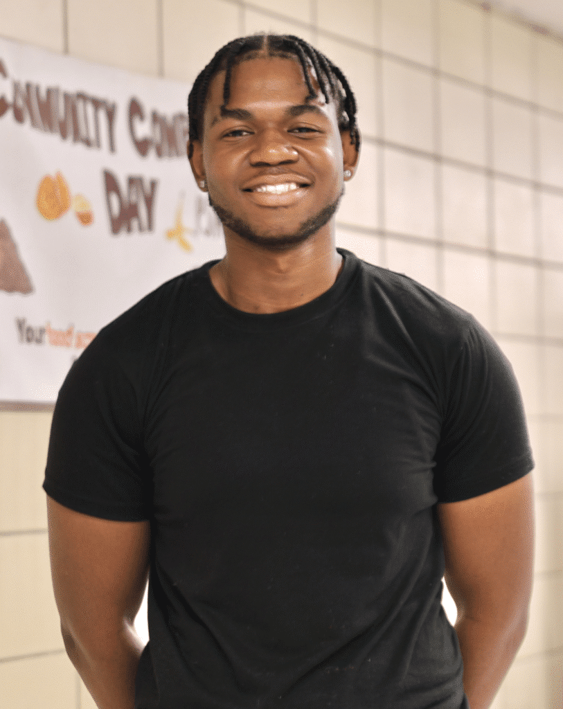 A young man in a black t-shirt and khaki pants stands smiling indoors near a tiled wall with a “Community Care Day” sign in the background.